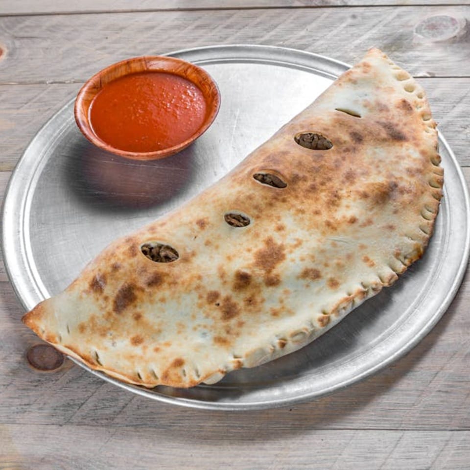 A golden-brown calzone with visible filling through vents, served with a side of red sauce on a metal tray.