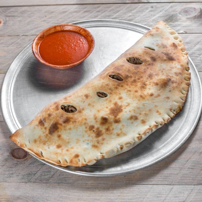 A golden-brown calzone with visible filling through vents, served with a side of red sauce on a metal tray.
