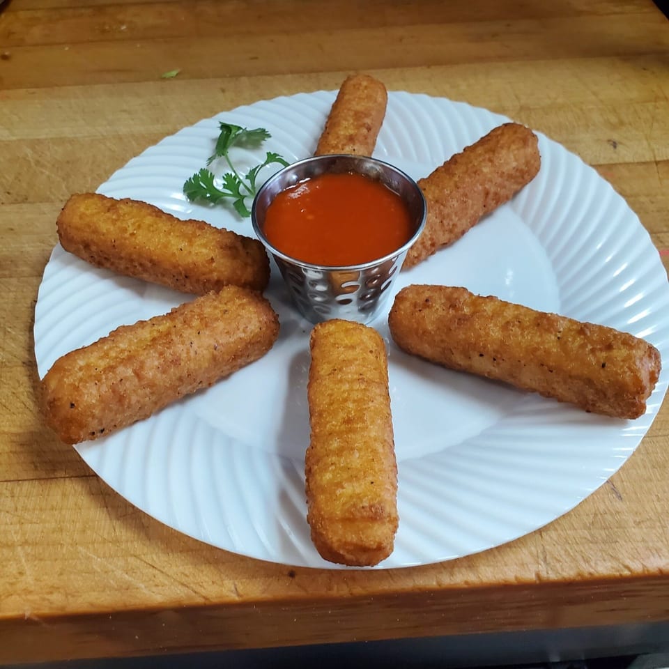 Six golden-brown mozzarella sticks arranged around a small metal cup of red marinara sauce on a white plate.