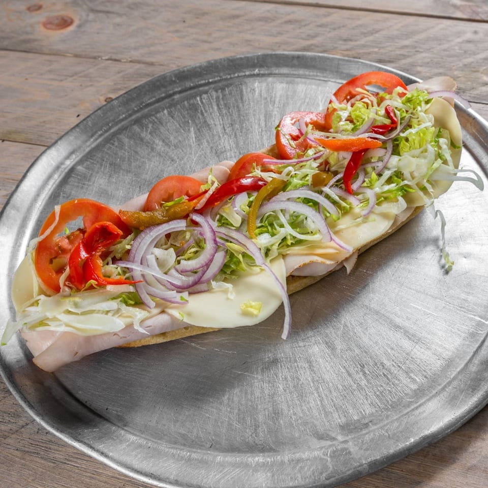 A large sub sandwich with turkey, cheese, lettuce, tomatoes, onions, and peppers on a metal tray.