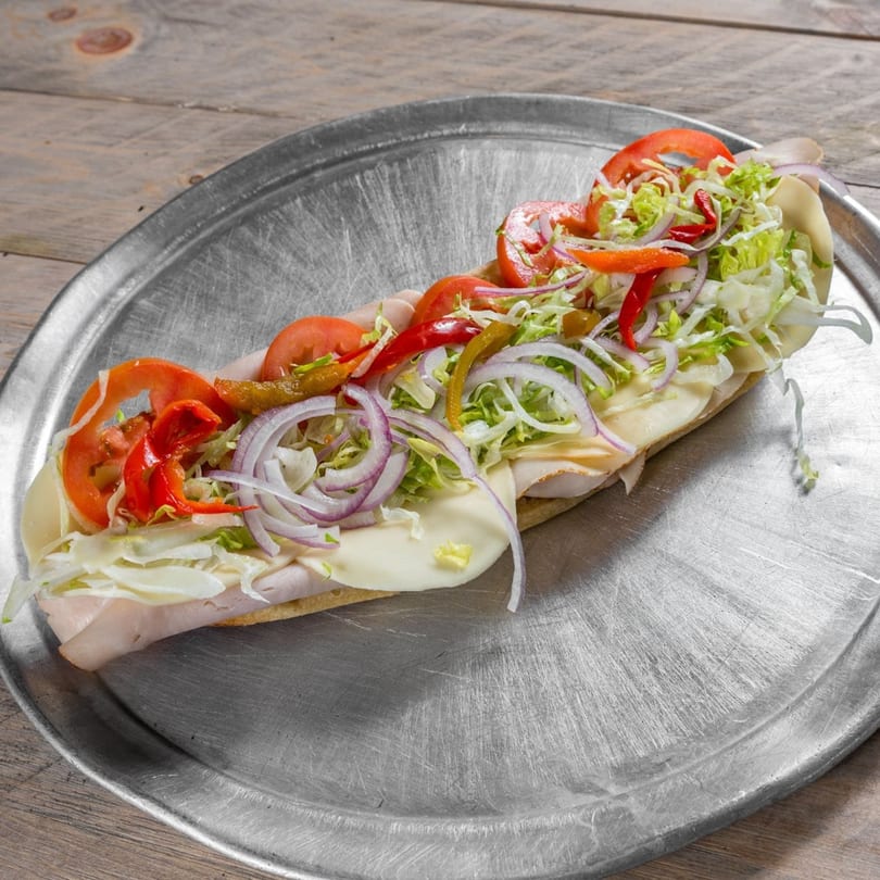 A large sub sandwich with turkey, cheese, lettuce, tomatoes, onions, and peppers on a metal tray.