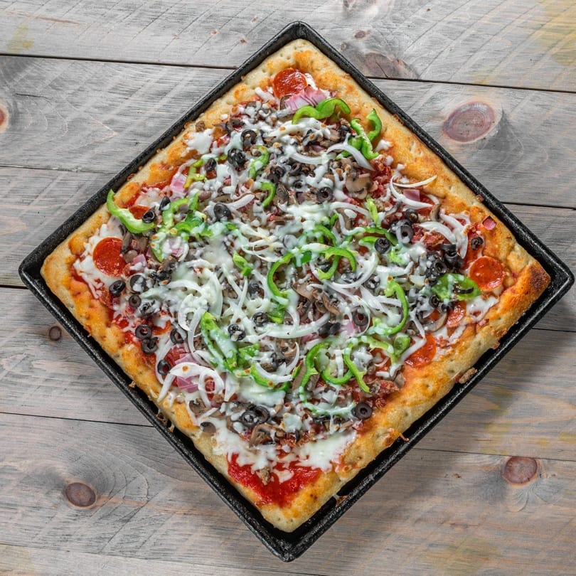 Square Detroit-style pizza with pepperoni, olives, green peppers, onions, and mushrooms on a wooden table.
